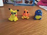 L'image montre trois figurines en pâte à modeler. À gauche, il y a un Pikachu jaune avec des joues rouges. Au milieu, un Charmander orange, assis. À droite, un Squirtle bleu, également assis. Les trois figurines sont bien colorées et semblent être des personnages inspirés de la franchise Pokémon. Elles sont placées sur une surface en bois.