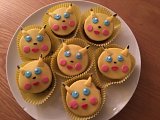 L'image montre un plateau de cupcakes décorés pour ressembler à Pikachu, un personnage célèbre de l'univers Pokémon. Les cupcakes ont un glaçage jaune avec des yeux en sucre bleu, des joues roses et une petite bouche. Ils ont également des petites oreilles pointues qui sortent du glaçage, renforçant leur ressemblance avec le personnage. Ils sont présentés dans des caissettes en papier de couleur jaune sur une assiette blanche.