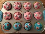 L'image montre un plateau de muffins décorés de manière ludique. Il y a douze muffins, chacun ornés de glaçage coloré. Les six muffins du haut sont recouverts d'un glaçage rose et représentent des visages amusants, avec des yeux en sucre blancs et des bouches rouges. Les six muffins du bas affichent des glaçages bleus et verts, également avec des visages souriants, mais avec des motifs et des couleurs différentes. L'ensemble donne une impression joyeuse et créative, parfait pour une fête ou une occasion particulière.