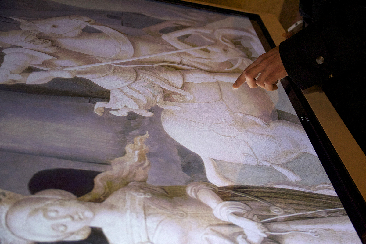L'image montre une personne pointant du doigt une œuvre d'art affichée sur un écran. L'œuvre représente une scène détaillée, probablement historique ou religieuse, avec des figures équestres et des personnages. Les couleurs sont délicates et les détails sont fins, suggérant un style classique. La personne interagit avec l'image, attirant l'attention sur un détail spécifique de la composition.