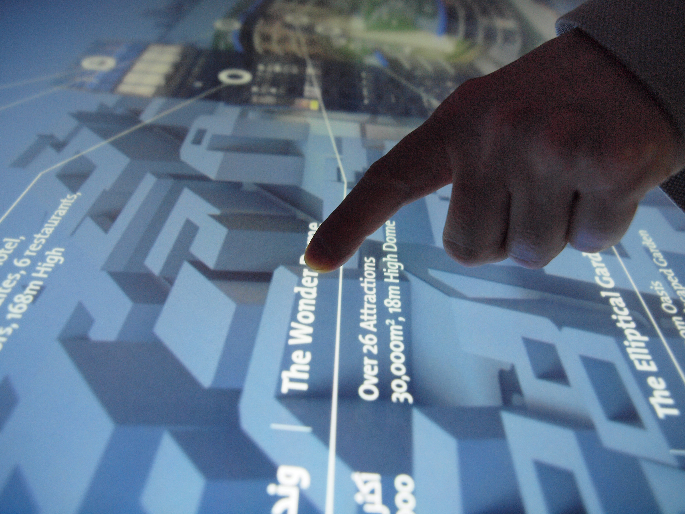 L'image montre une main pointant un écran interactif, probablement un plan ou une carte. On peut voir des informations sur des attractions, avec des mots en français et en arabe. Le texte indique "The Wonder" et mentionne plus de 26 attractions avec des détails sur leur taille. L'écran semble être une interface numérique conçue pour guider les visiteurs.
