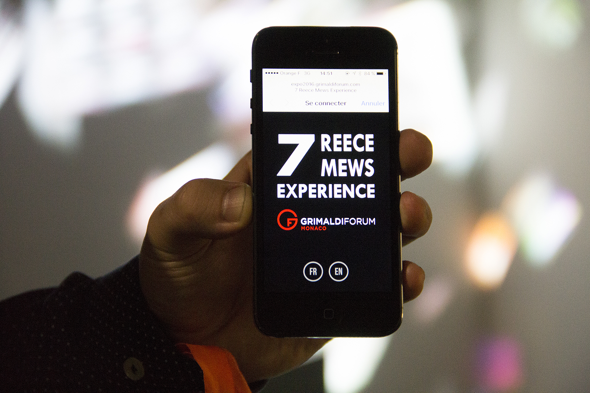 L'image montre un téléphone portable tenu en main, affichant un écran avec des informations sur un événement. Le texte en gros caractères mentionne "7 REECE MEWS EXPERIENCE", et un logo avec le nom "GRIMALDI FORUM MONACO" est visible. Il y a également des options pour se connecter, ainsi que des indications pour choisir la langue (FR pour français et EN pour anglais). L'arrière-plan est flou, avec des formes colorées.