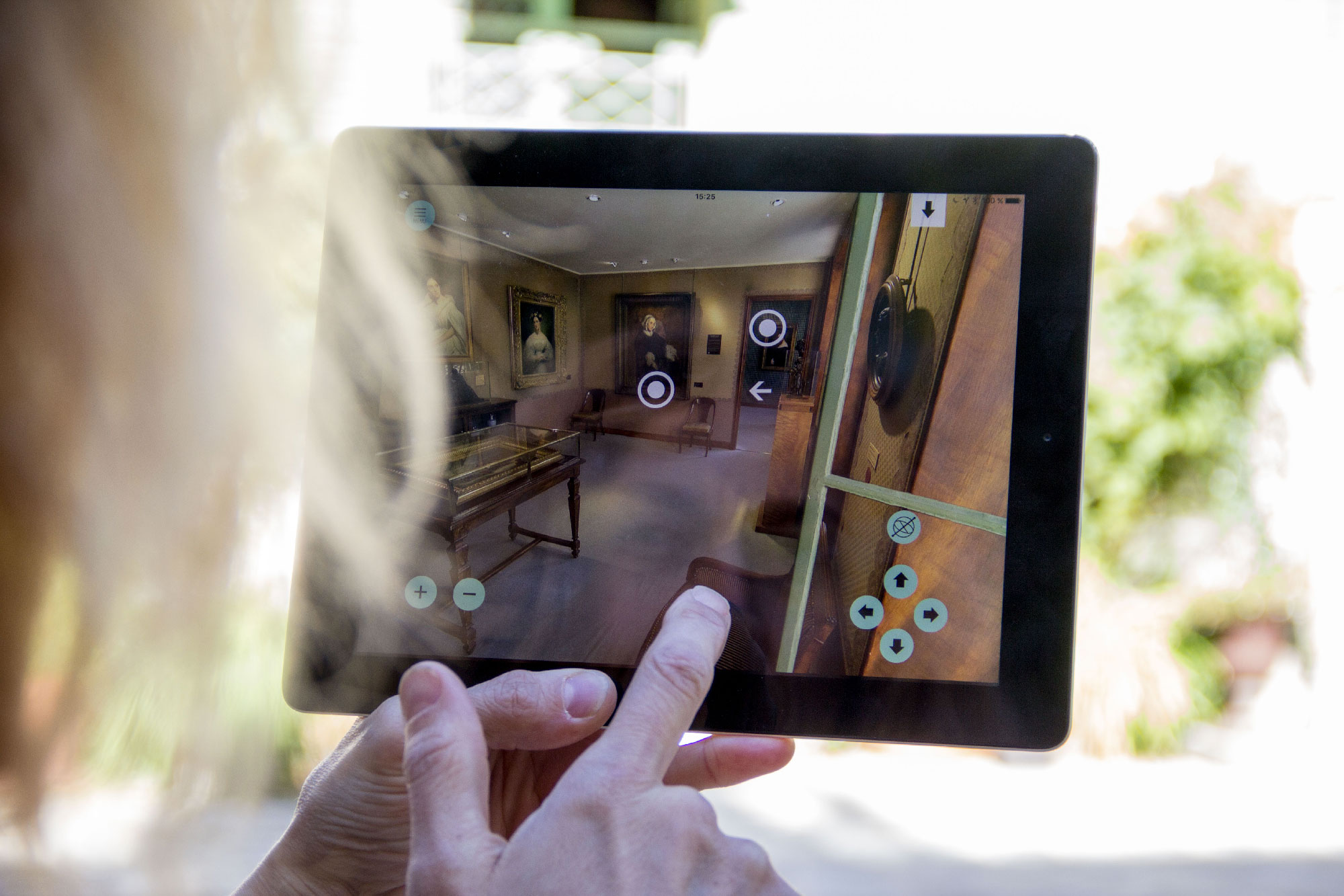 L'image montre une personne tenant une tablette dans un espace intérieur. Sur l'écran de la tablette, on peut voir une représentation en 3D d'une pièce, probablement issue d'une visite virtuelle. Des éléments de navigation, comme des cercles et des flèches, sont visibles sur l'écran, suggérant que l'utilisateur peut interagir avec l'environnement virtuel. L'arrière-plan de la photo montre une zone bien éclairée, probablement un jardin ou un patio, ce qui donne une ambiance agréable à la scène.