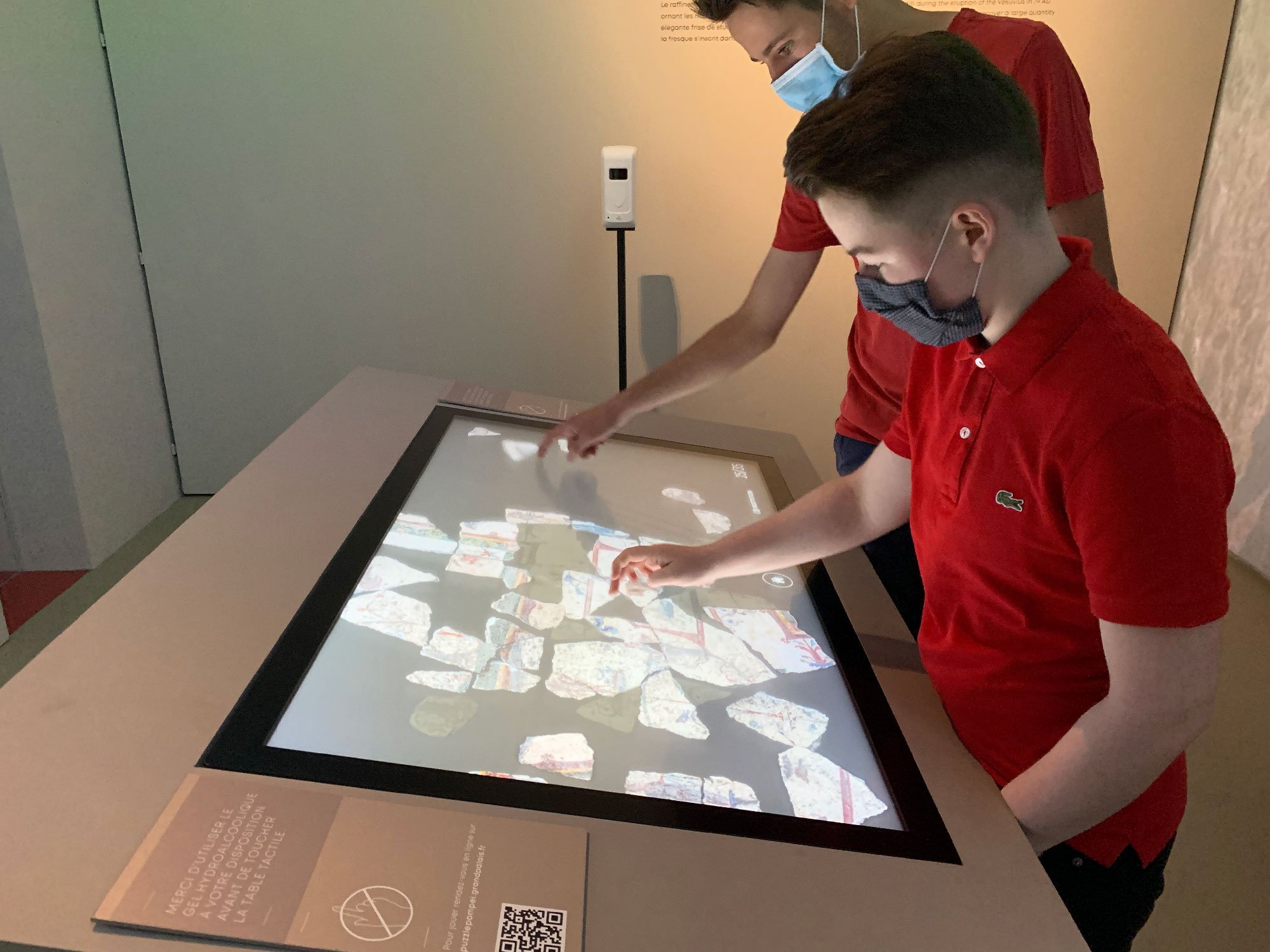 L'image montre deux jeunes garçons interactant avec un écran tactile en exposant des éléments. Ils portent des masques en raison de mesures sanitaires. L'écran affiche de nombreux morceaux colorés, peut-être des artefacts ou des éléments informatiques, et les garçons semblent manipuler ces objets virtuels. L'environnement est moderne et éducatif, suggérant qu'ils se trouvent dans un musée ou une exposition.
