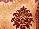 L'image montre un coussin décoratif avec un motif riche et orné. Le fond est principalement de couleur crème, tandis que le motif central est en rouge foncé, entouré de détails aux teintes dorées et ocre. Les motifs évoquent des motifs floraux ou baroques, ajoutant une touche élégante et sophistiquée à l'ensemble du coussin. Le design semble complexe, soulignant un style classique.