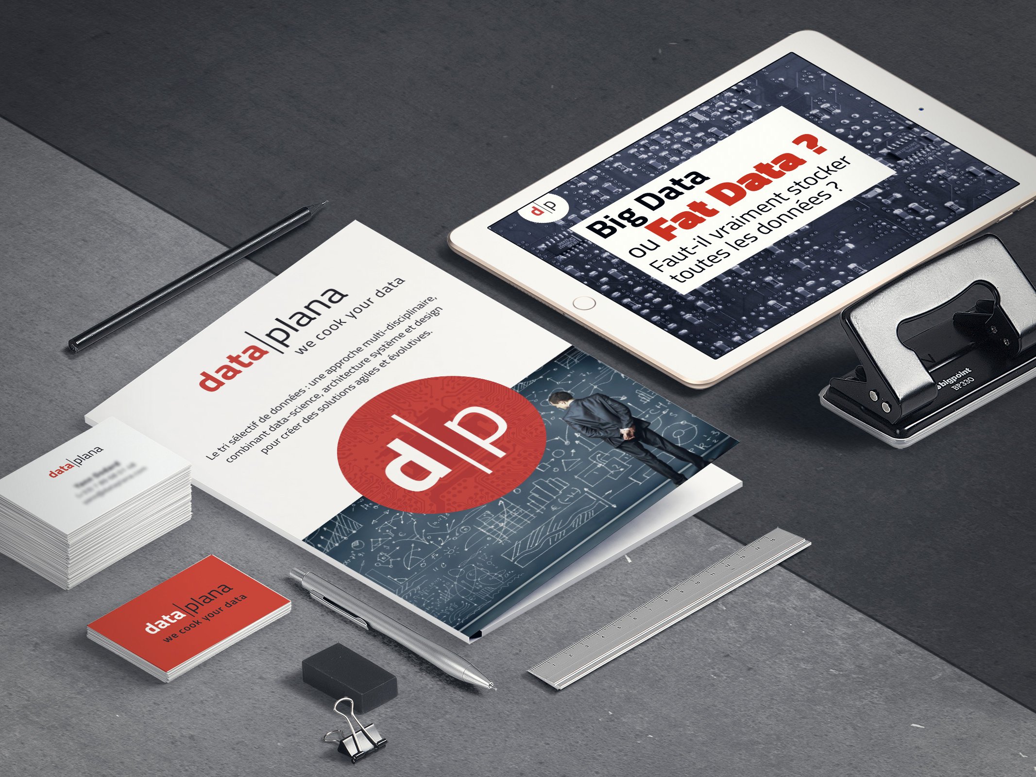Cette image présente un bureau moderne et bien organisé. Au centre, il y a un document intitulé "data|plana" avec un design graphique, entouré de divers objets de bureau. On voit également une tablette numérique affichant des informations sur le Big Data. À côté, il y a des cartes de visite, un stylo et un clip, le tout sur un fond en béton. L'ensemble dégage une ambiance professionnelle et technologique, axée sur la gestion des données.