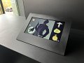 L'image montre un écran tactile placé sur une surface noire. L'écran présente des illustrations, dont une silhouette d'homme en costume avec une main posée, ainsi que plusieurs objets qui semblent être des artefacts ou bijoux, comme une montre de poche et une bague. Le fond de l'écran est sobre, mettant en valeur les éléments graphiques. On peut apercevoir à l'extérieur à travers une fenêtre, suggérant que l'image a été prise dans un espace d'exposition ou un musée.