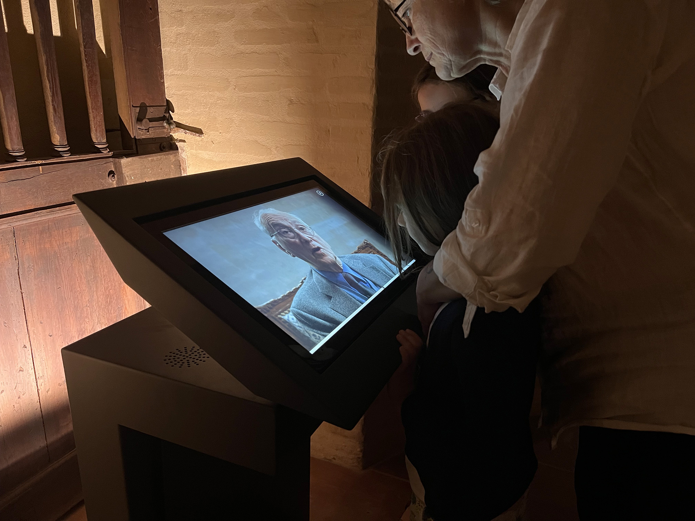L'image montre une interaction entre deux enfants et un adulte devant un écran interactif. L'écran affiche un homme qui semble s'adresser à eux, peut-être en partageant une histoire ou une explication. L'environnement semble être à l'intérieur d'un bâtiment ancien, avec des murs en pierre et une porte en bois en arrière-plan. Les enfants se tiennent près de l'écran, captivés par ce qu'ils voient. L'adulte, qui est légèrement penché en avant, semble également intéressé par le contenu diffusé.