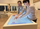L'image montre deux personnes, un homme et une femme, qui interagissent avec une table tactile. Ils semblent analyser ou manipuler des éléments sur l'écran avec leurs doigts, tandis qu'en arrière-plan, on peut apercevoir des sculptures ou des œuvres exposées dans un musée. L'espace est lumineux et bien agencé, mettant en valeur les objets sur display.