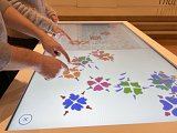 L'image montre des mains de plusieurs personnes posées sur un écran tactile interactif. Les doigts pointent vers des motifs de fleurs colorés affichés à l'écran, qui comprennent des teintes de bleu, orange et d'autres couleurs. L'environnement a l'air d'un musée ou d'une exposition, où les visiteurs interagissent avec une œuvre numérique ou un jeu éducatif.
