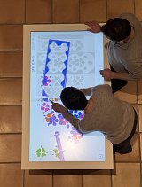 L'image montre deux personnes interagissant avec une table numérique. Ils semblent s'engager dans une activité créative, utilisant leurs mains pour manipuler des éléments colorés affichés sur l'écran. Les motifs évoquent des fleurs et des formes variées, et il y a un cadre bleu qui semble délimiter une zone d'intérêt. L'atmosphère est collaborative et ludique, mettant en avant l'interaction avec la technologie.