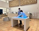 L'image montre deux personnes qui interagissent avec un écran tactile dans un espace intérieur. Ils semblent concentrés sur ce qu'ils font, et l'éclairage de la table crée un effet visuel attrayant. L'environnement est moderne, avec des murs décorés de motifs et des œuvres d'art. Le sol est en carrelage, et il y a des éléments décoratifs en arrière-plan, comme des sculptures ou des installations artistiques.