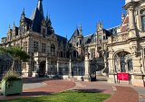 L'image montre un imposant bâtiment architectural de style classique avec des éléments gothiques. La façade est richement décorée, avec des balcons et des sculptures. On y aperçoit des toits pointus, des fenêtres ornées et des détails artistiques. Au premier plan, il y a un parterre verdoyant avec des fleurs, et une allée pavée mène à l'entrée du bâtiment, qui est protégée par une grille élégante. Le ciel est bleu et ensoleillé, ajoutant une ambiance lumineuse à la scène.