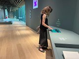 L'image montre une personne debout dans un musée ou une galerie d'art. Elle est vêtue d'un top noir et d'un pantalon long, et elle interagit avec un écran tactile placé sur une table. Le décor environnant est moderne, avec un sol en bois clair et des murs gris. On aperçoit également d'autres œuvres d'art et objets d'exposition dans l'espace. La lumière naturelle semble entrer par des fenêtres, créant une ambiance agréable.