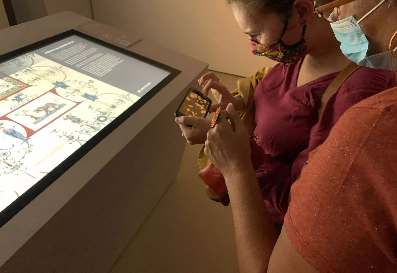 L'image montre deux personnes observant un écran interactif dans un musée. Elles semblent être en train d'explorer des contenus sur une tablette ou un téléphone. L'une des personnes est vêtue d'un haut de couleur bordeaux et porte un masque. L'autre personne, à côté, tient un accessoire ou un petit objet. L'éclairage de la pièce est doux, créant une atmosphère calme et contemplative. Les deux personnes semblent concentrées sur leur activité.