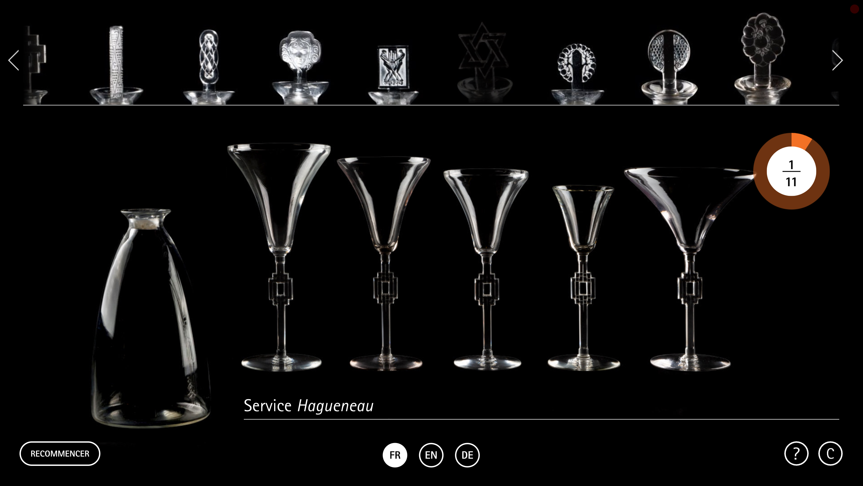 L'image présente un ensemble de verres et de couverts en cristal, disposés sur un fond noir. On peut voir plusieurs types de verres, notamment des flûtes et des coupes, ainsi qu'une carafe de forme élégante. Les pièces sont ornées de motifs raffinés, caractérisant un style artistique classique. Cet ensemble est identifié comme étant un "Service Haguenau". La présentation met en valeur les détails du design et la brillance du cristal.