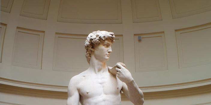Cette image représente une sculpture célèbre, le "David" de Michel-Ange. La statue est en marbre et dépeint un jeune homme nu, d'une beauté classique, avec des détails anatomiques précis. Il se tient debout, tourné légèrement sur le côté, avec un regard déterminé. Sa main gauche repose sur son épaule, tandis que son bras droit est le long de son corps. Le fond de la pièce est sobre, mettant en valeur la statue au centre.