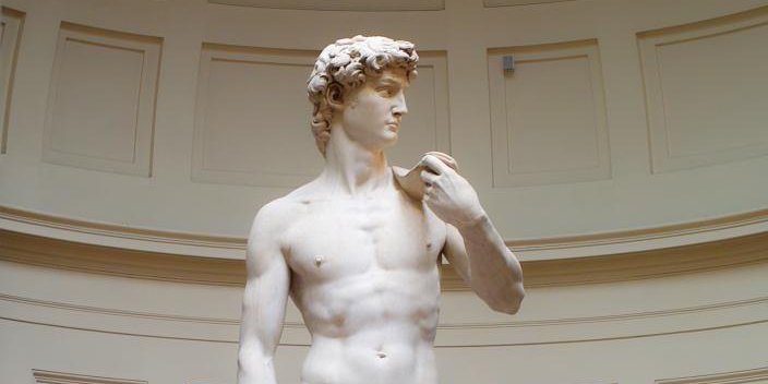 L'image représente une célèbre sculpture en marbre, connue sous le nom de David, réalisée par Michel-Ange. La statue montre un jeune homme, nu, avec une posture dynamique. Il est tourné légèrement sur le côté, regardant vers l'horizon, affichant une expression de concentration. Les détails anatomiques sont précis, mettant en valeur la musculature et le réalisme de la figure. L'arrière-plan est sobre, ce qui met en valeur la statue elle-même.