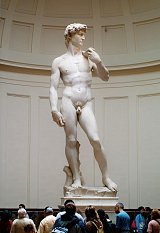 L'image montre la célèbre sculpture du David de Michel-Ange, qui est exposée dans un grand espace intérieur. La statue, réalisée en marbre, représente un jeune homme nu avec une posture gracieuse et confiante. Il est debout, légèrement tourné, avec un bras replié et l'autre reposant à ses côtés. Le visage de la statue exprime une concentration intense, et son corps est finement détaillé, mettant en valeur l'anatomie humaine. Autour de la sculpture, un groupe de visiteurs semble admirer l'œuvre. Les murs de la salle sont clairs et épurés, mettant en avant la beauté de la statue elle-même.