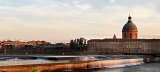 Cette image montre une vue paisible d'une ville au bord de l'eau. On peut apercevoir un pont élégant, des bâtiments historiques avec des façades en briques, et un dôme majestueux en arrière-plan. L'eau s'écoule doucement avec des petites chutes, tandis que la lumière du soleil crée une ambiance chaleureuse, suggérant un moment agréable, probablement au crépuscule. Le paysage semble harmonieux, avec des arbres verdoyants ajoutant une touche de nature à la scène urbaine.