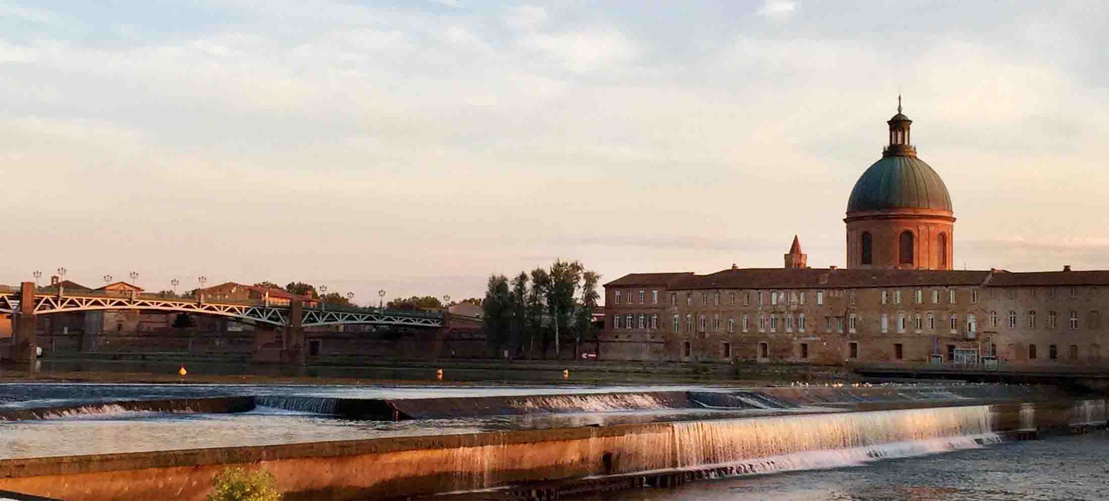 Cette image montre une vue paisible d'une ville au bord de l'eau. On peut apercevoir un pont élégant, des bâtiments historiques avec des façades en briques, et un dôme majestueux en arrière-plan. L'eau s'écoule doucement avec des petites chutes, tandis que la lumière du soleil crée une ambiance chaleureuse, suggérant un moment agréable, probablement au crépuscule. Le paysage semble harmonieux, avec des arbres verdoyants ajoutant une touche de nature à la scène urbaine.