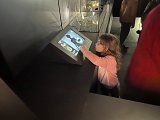 L'image montre une petite fille en train d'interagir avec un écran tactile, probablement dans un musée. Elle est concentrée, touchant l'écran avec un doigt. En arrière-plan, on peut voir des vitrines exposant des objets d'art ou des artefacts. D'autres visiteurs sont présents, mais leur présentation est floue, mettant l'accent sur l'enfant. La lumière est tamisée, créant une atmosphère muséale.