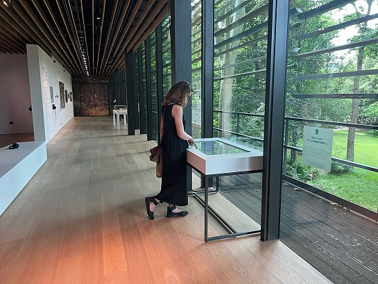 L'image montre une personne se tenant devant une vitrine dans un espace d'exposition. Elle examine quelque chose à l'intérieur de la vitrine, qui est éclairée par la lumière naturelle qui entre par de grandes fenêtres. On peut voir un paysage verdoyant à l'extérieur, suggérant que l'espace est entouré de nature. Le décor intérieur est moderne, avec des murs blancs et un sol en bois. La personne porte une robe noire et semble plongée dans sa contemplation.
