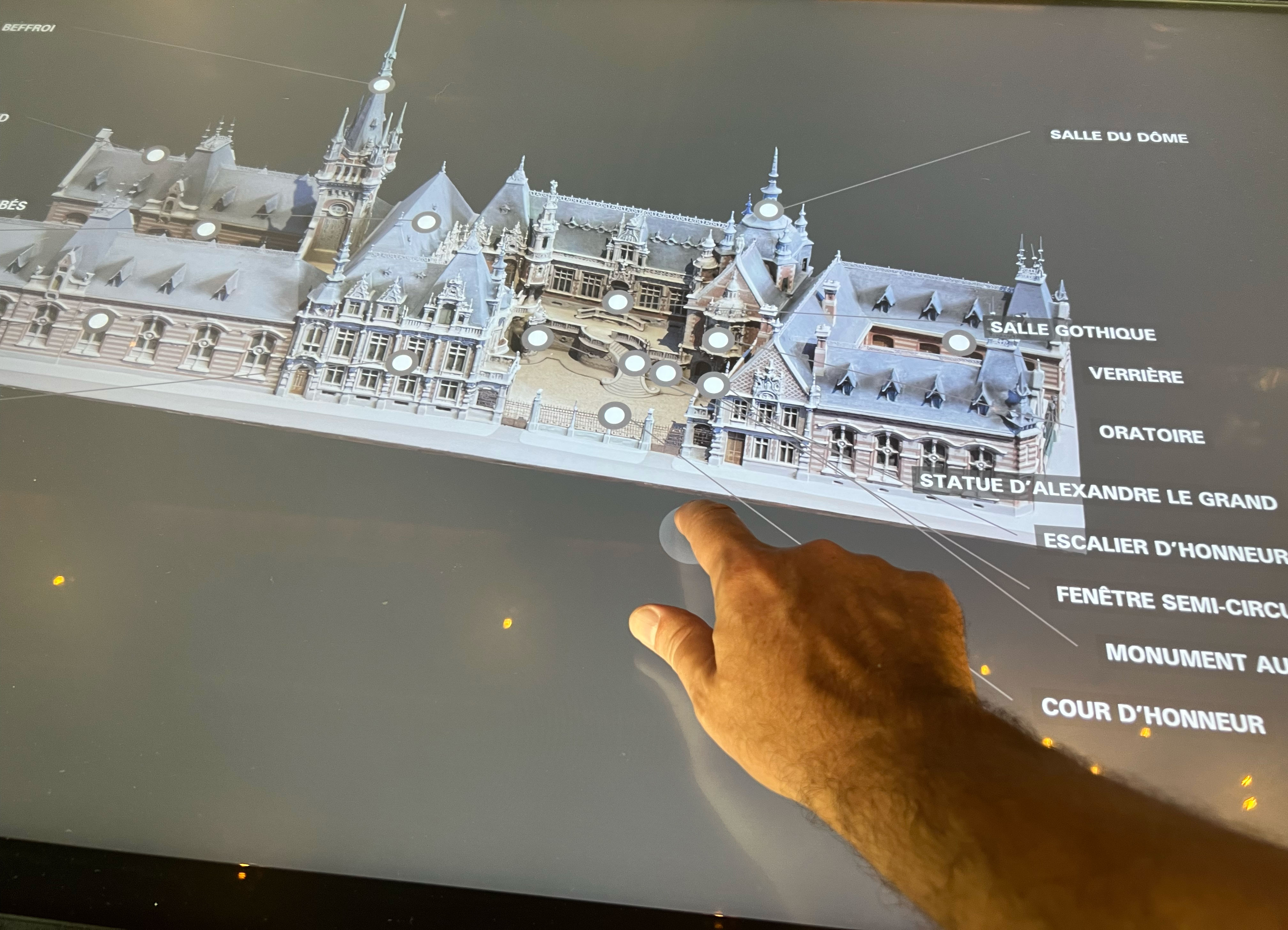 L'image montre un plan interactif d'un bâtiment architectural, probablement un hôtel de ville ou un monument historique. Une main tactile pointe vers une partie du bâtiment, indiquant une salle ou une section spécifique. Le texte autour de l'image mentionne divers éléments architecturaux, comme la "Salle du Dôme," la "Salle Gothique," et la "Statue d'Alexandre le Grand," suggérant que ce lieu a une riche histoire et plusieurs zones d'intérêt.