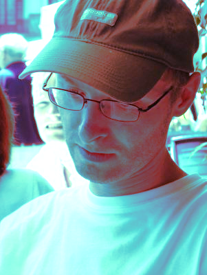 L'image montre un homme portant une casquette et des lunettes, concentré sur quelque chose qu'il lit ou consulte. On peut voir des personnes en arrière-plan, mais elles ne sont pas clairement identifiables. L'éclairage donne à la scène une teinte légèrement colorée, créant une ambiance particulière.