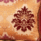 L'image montre un coussin décoratif avec un motif riche et orné. Le fond est principalement de couleur crème, tandis que le motif central est en rouge foncé, entouré de détails aux teintes dorées et ocre. Les motifs évoquent des motifs floraux ou baroques, ajoutant une touche élégante et sophistiquée à l'ensemble du coussin. Le design semble complexe, soulignant un style classique.