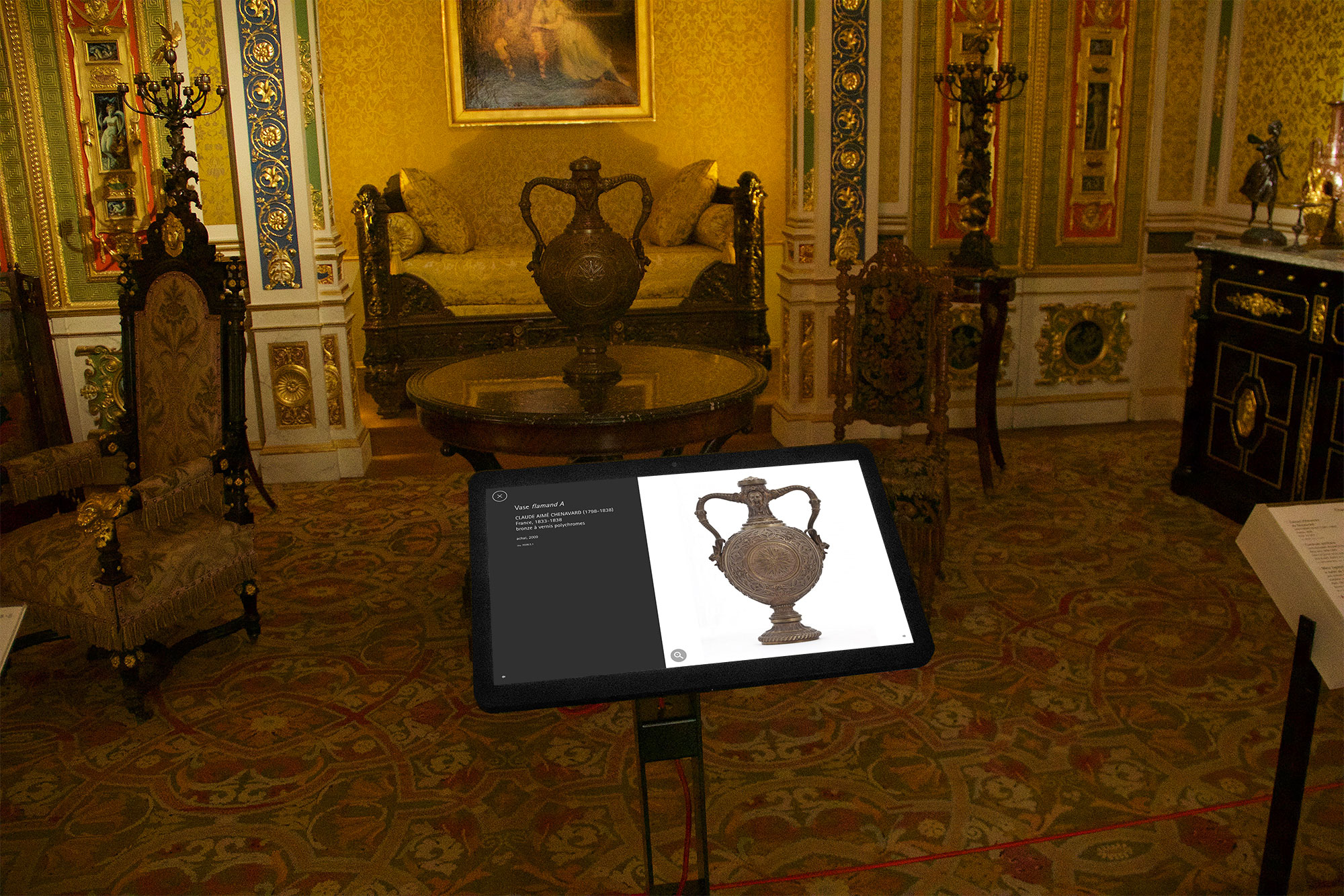 L'image montre une pièce richement décorée, probablement dans un musée ou un palais, avec des détails architecturaux élaborés. Au centre, il y a une table sur laquelle repose une tablette affichant une représentation d'une belle urne ou vase ornée. En arrière-plan, on aperçoit un canapé et des chaises élégantes, toutes avec des motifs raffinés. Le sol est recouvert d'un tapis orné aux couleurs vives. Les murs sont décorés avec des motifs complexes et des œuvres d'art. L'ensemble dégage une atmosphère historique et artistique.