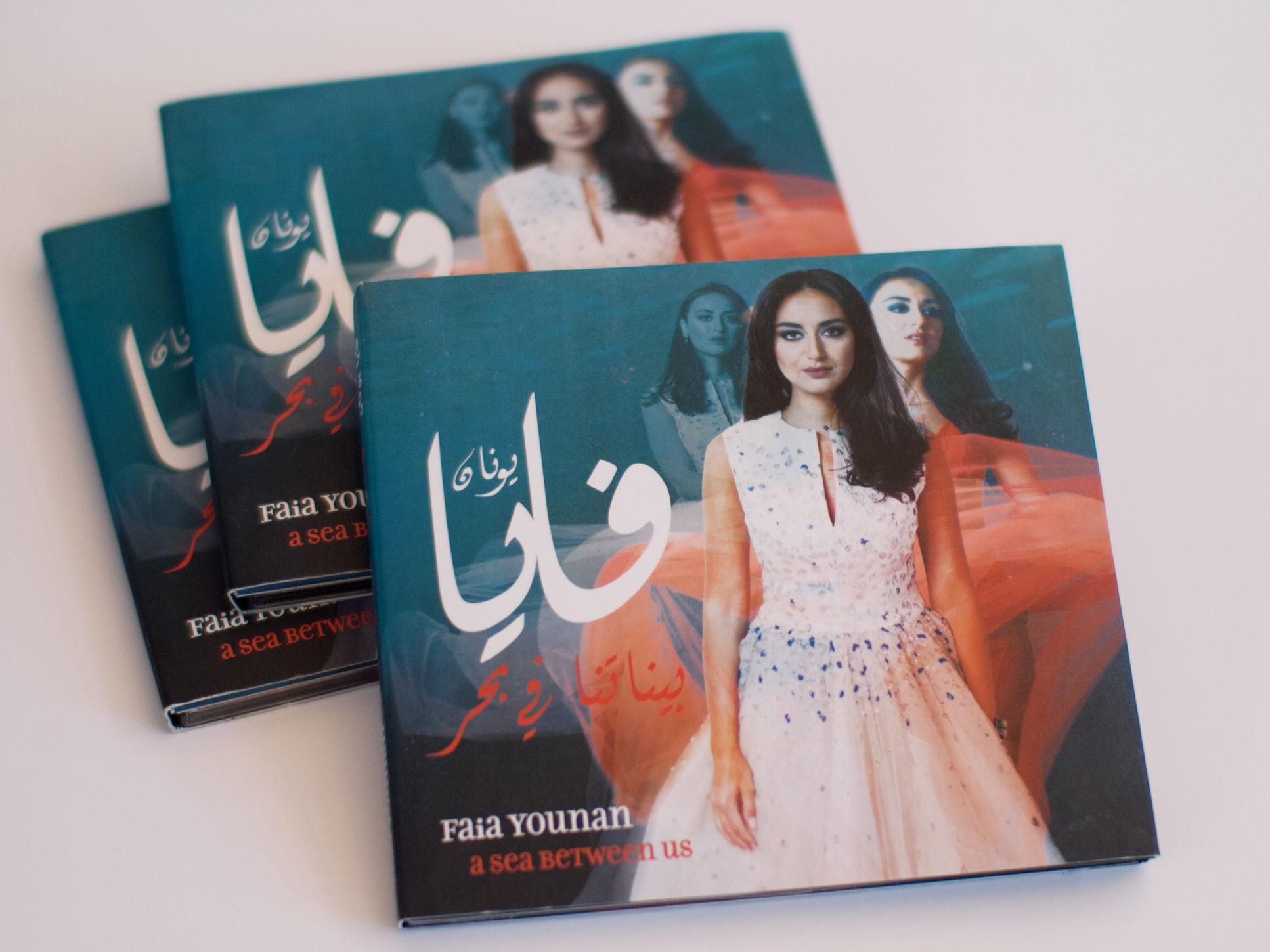 L'image montre plusieurs exemplaires d'un album musical au design coloré. La couverture présente une femme au regard captivant, habillée d'une robe élégante, qui se trouve devant un arrière-plan artistique. Le titre de l'album, écrit en arabe et en anglais, est visible en haut et en bas de l'image. Les couleurs dominantes sont une combinaison de teintes chaudes et froides, donnant une atmosphère vivante et moderne à l'ensemble de la présentation.