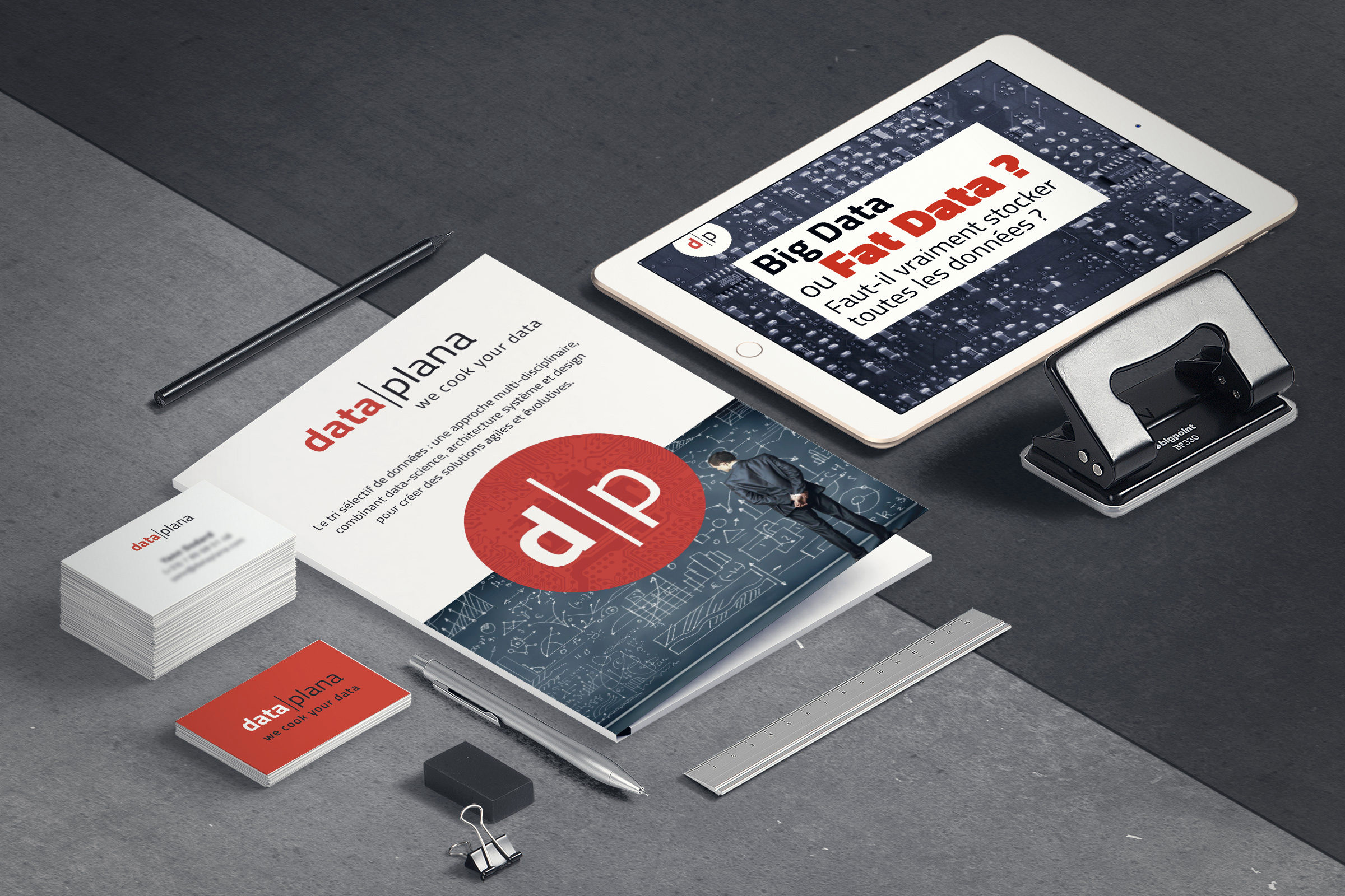 Cette image présente un bureau moderne et bien organisé. Au centre, il y a un document intitulé "data|plana" avec un design graphique, entouré de divers objets de bureau. On voit également une tablette numérique affichant des informations sur le Big Data. À côté, il y a des cartes de visite, un stylo et un clip, le tout sur un fond en béton. L'ensemble dégage une ambiance professionnelle et technologique, axée sur la gestion des données.