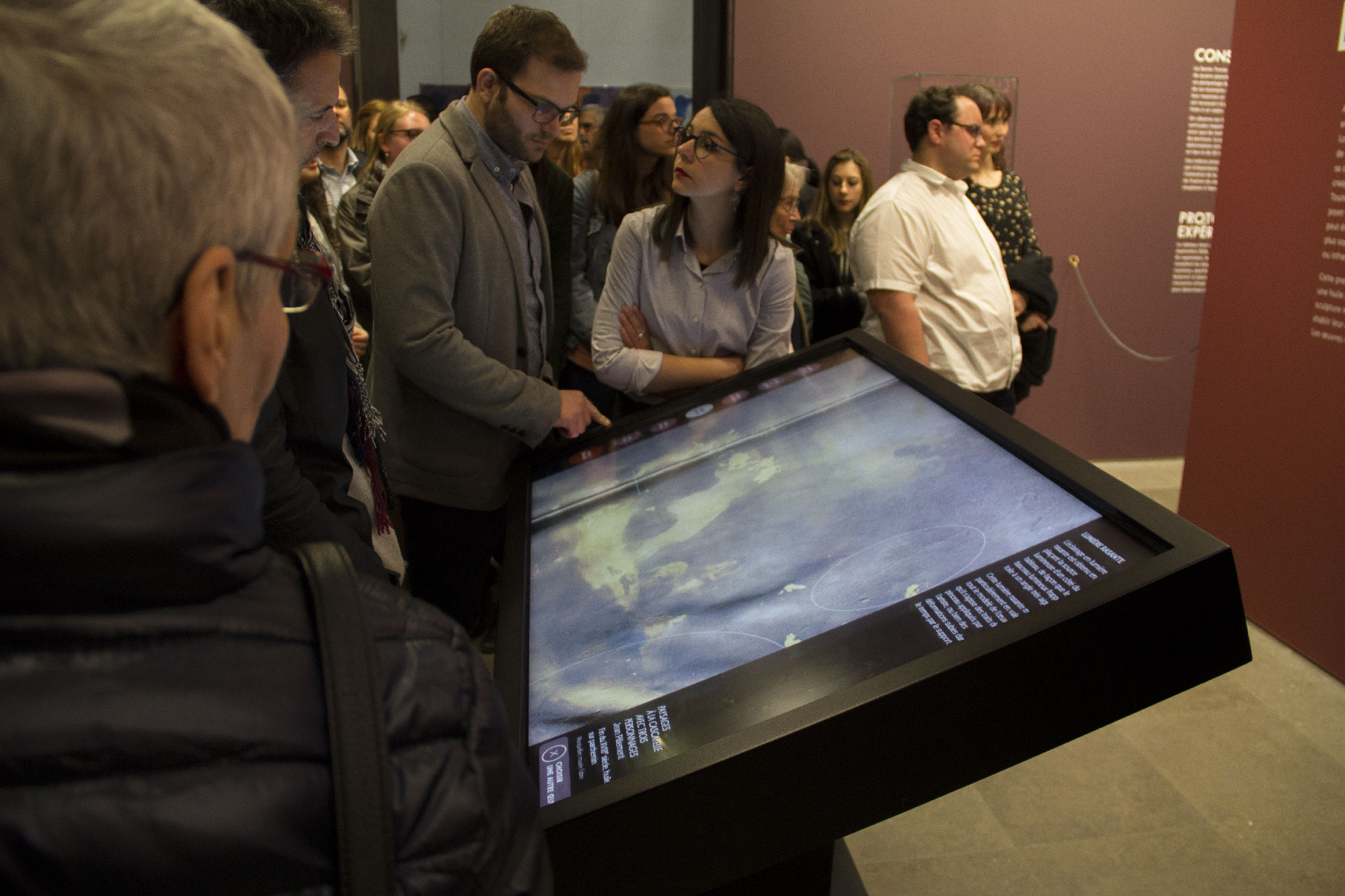 L'image montre un groupe de personnes rassemblées autour d'un grand écran tactile interactif. Deux personnes, probablement un homme et une femme, semblent particulièrement intéressées par ce qui est affiché à l'écran. L'arrière-plan est flou, mais on peut voir d'autres visiteurs curieux observant l'exposition. Le décor semble être celui d'un musée ou d'une galerie, avec des murs d'une couleur sombre.
