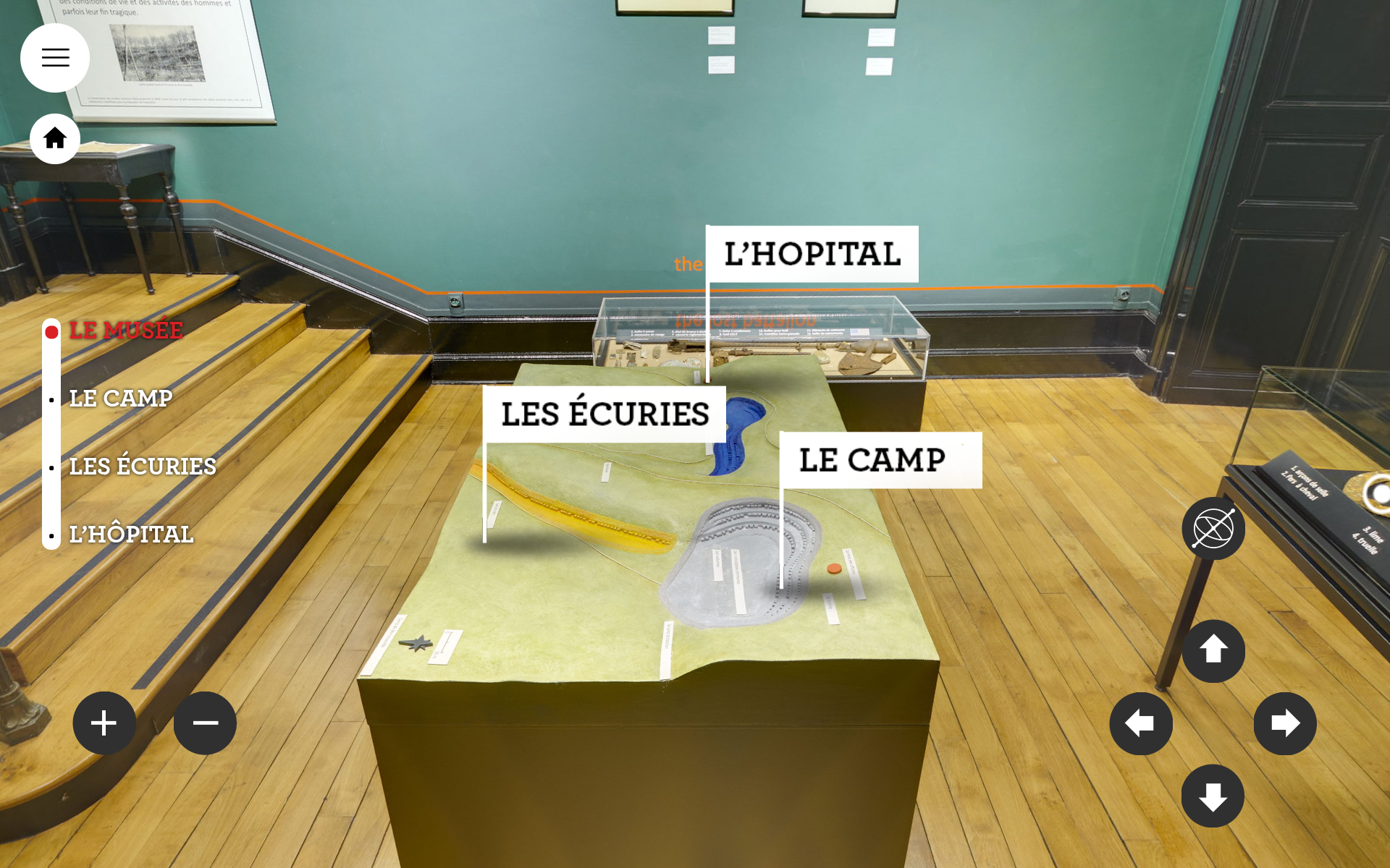 L'image montre une exposition dans un musée. Sur une table d'exposition, plusieurs éléments de présentation sont étiquetés. On y trouve les termes « l'Hôpital », « les Écuries » et « le Camp ». L'environnement est élégant, avec des murs de couleur verte et un sol en bois. Des objets ou maquettes semblent être présentés sous une vitrine, suggérant une thématique historique.
