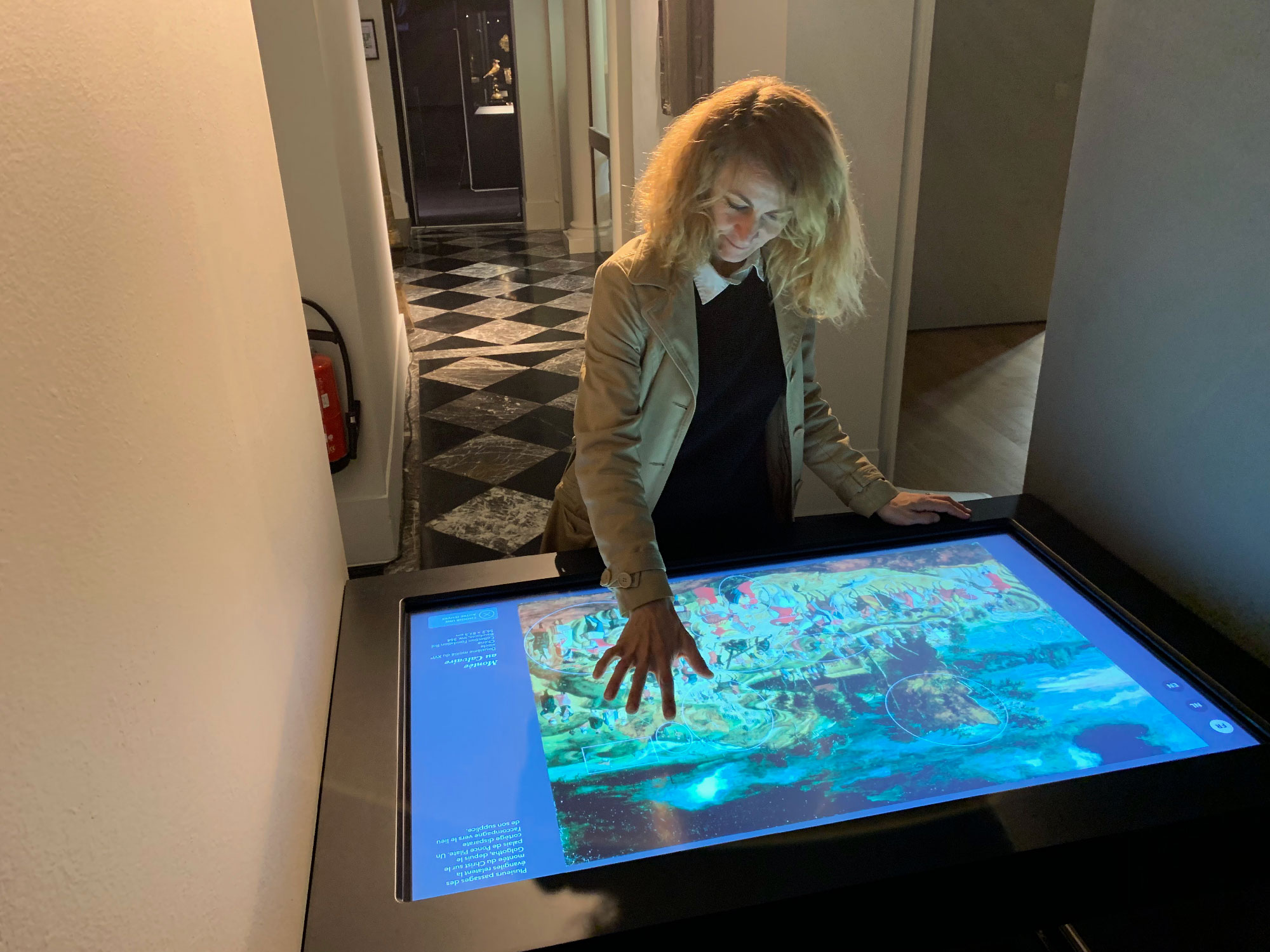 L'image montre une femme interagissant avec un écran tactile, probablement dans un musée ou une exposition. Elle semble intéressée par une œuvre d'art affichée sur l'écran. Le cadre de l'image présente des murs clairs et un sol en damier noir et blanc, indiquant un espace moderne et esthétique. On peut également apercevoir des portes en arrière-plan, suggérant un environnement artistique.