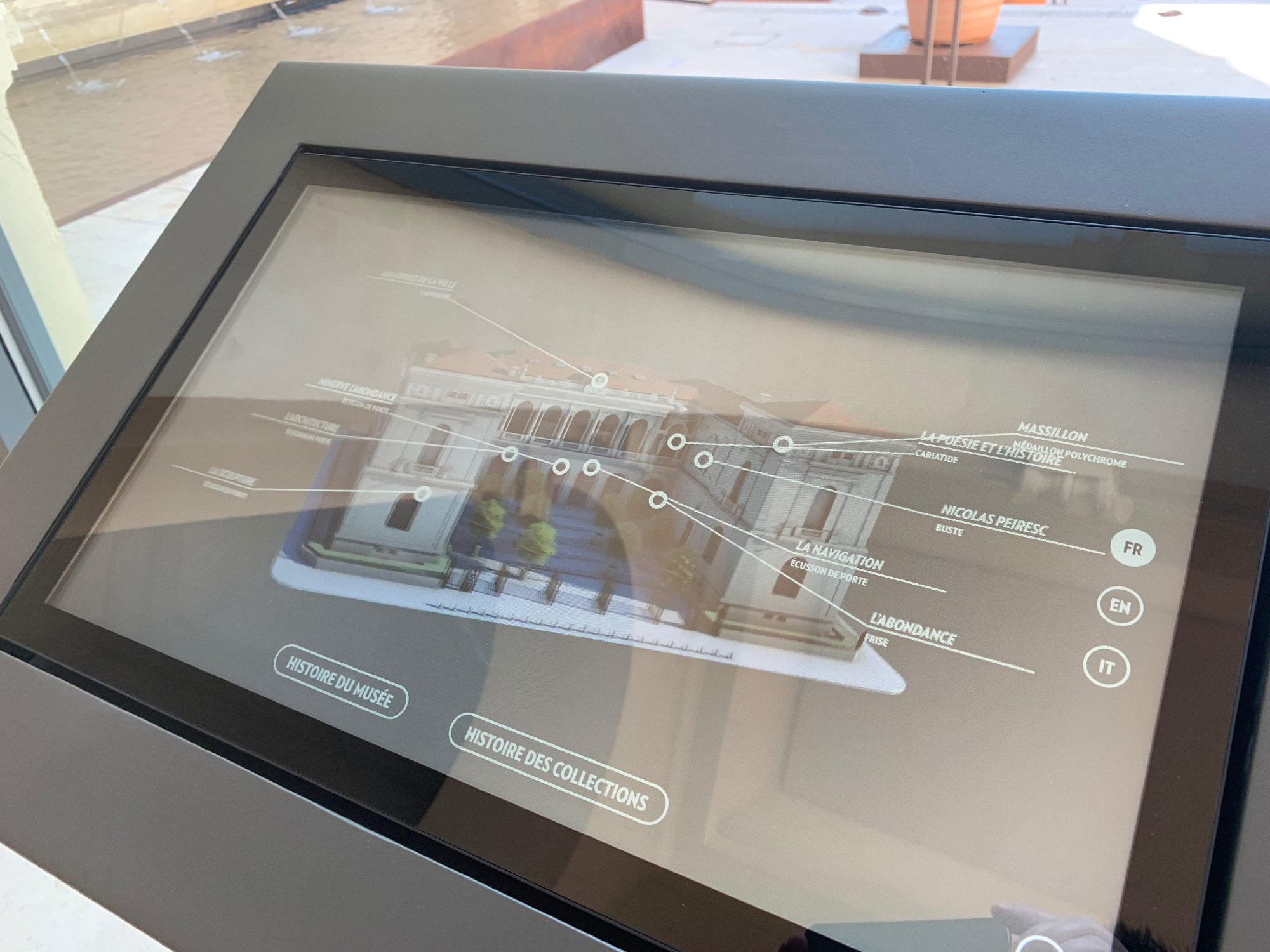 L'image montre un écran tactile interactif présentant une maquette ou une image en 3D d'un bâtiment, probablement un musée ou une galerie d'art. L'interface inclut des options de navigation en plusieurs langues, telles que le français (FR), l'anglais (EN) et l'italien (IT). Il y a des options pour explorer l'histoire du bâtiment ainsi que les collections qu'il abrite. En arrière-plan, on peut apercevoir un environnement extérieur, possiblement une terrasse ou un patio, avec de l'eau visible.