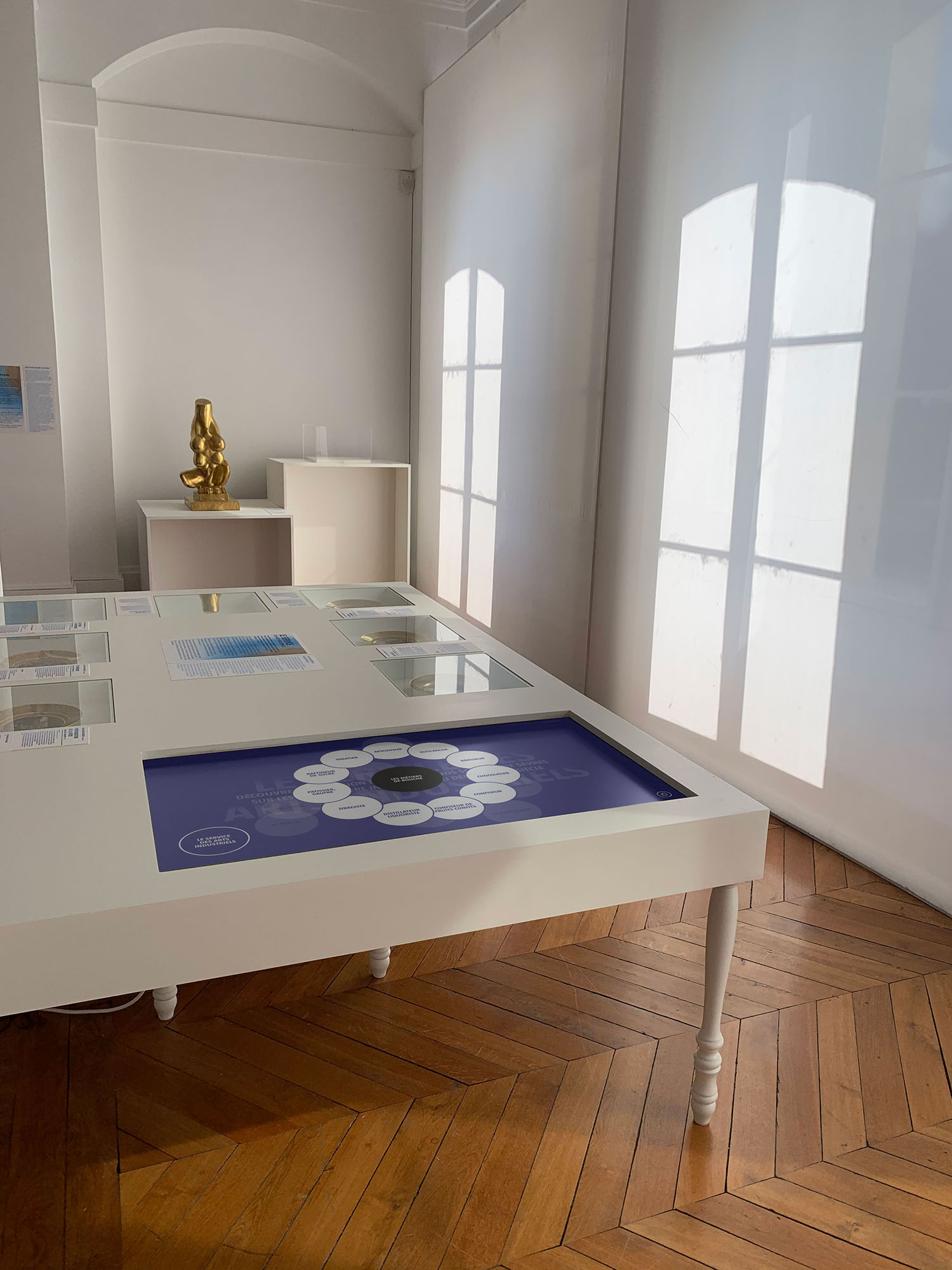 L'image montre une salle d'exposition avec un éclairage naturel provenant de fenêtres. Au centre, il y a une table blanche avec un écran tactile qui présente divers éléments visuels. Autour de cette table se trouvent des cadres avec des images ou des informations. À l'arrière-plan, il y a un objet doré sur une autre table, et l'ensemble de l'espace dégage une ambiance épurée et moderne grâce aux couleurs claires et au parquet en bois. Les ombres créées par la lumière filtrée ajoutent une touche esthétique à la scène.