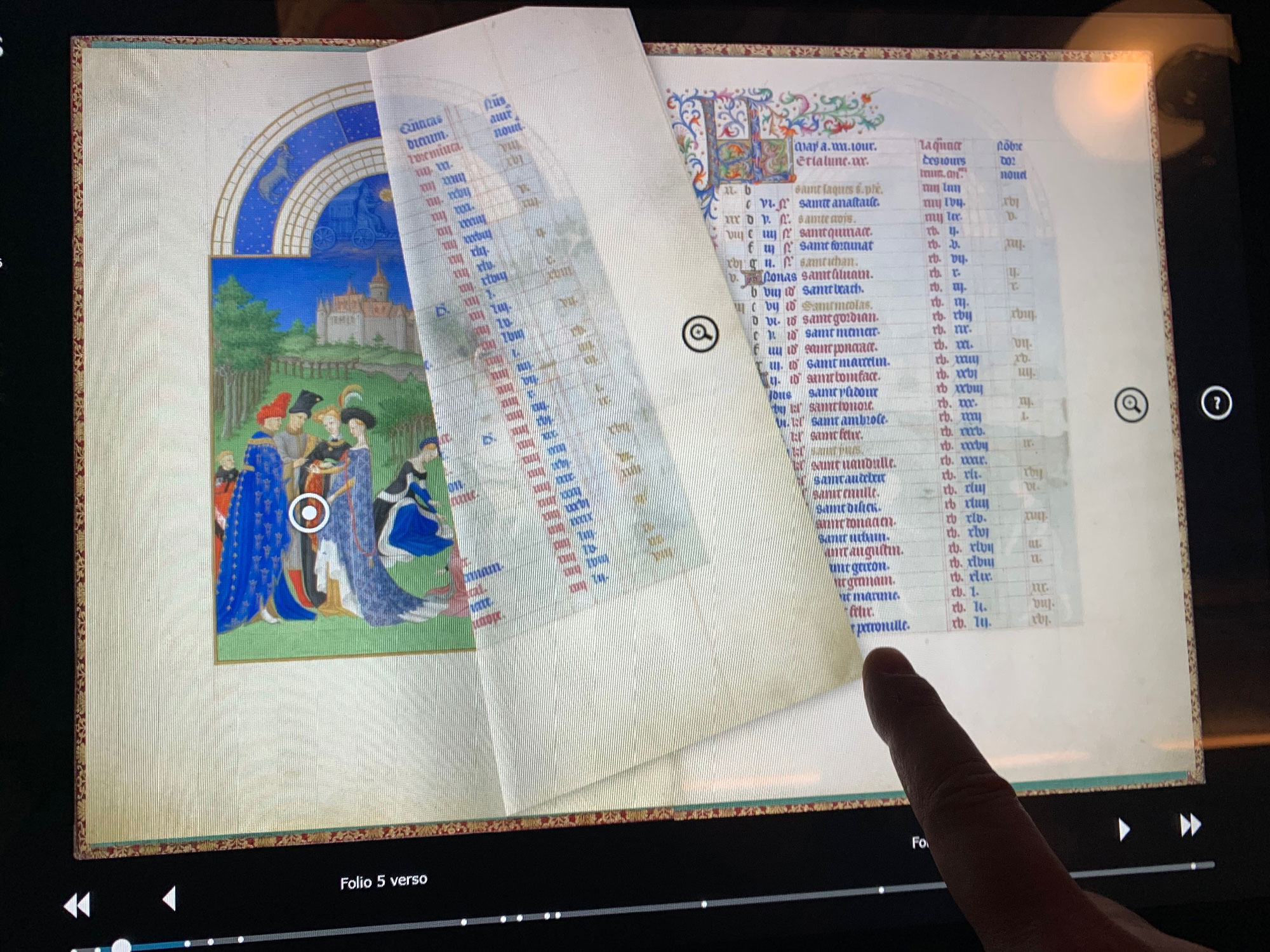 L'image montre une ancienne page manuscrite ouverte, probablement d'un livre d'heures ou d'un grimoire. À gauche, il y a une illustration colorée représentant des personnages vêtus de vêtements médiévaux, se tenant près d'un château. À droite, le texte est présenté sous forme de colonnes, avec des noms et des dates inscrits. Un doigt pointe vers l'une des pages, suggérant une analyse ou une attention particulière à un détail spécifique. Le décor est richement orné, reflétant le style artistique de l'époque médiévale.