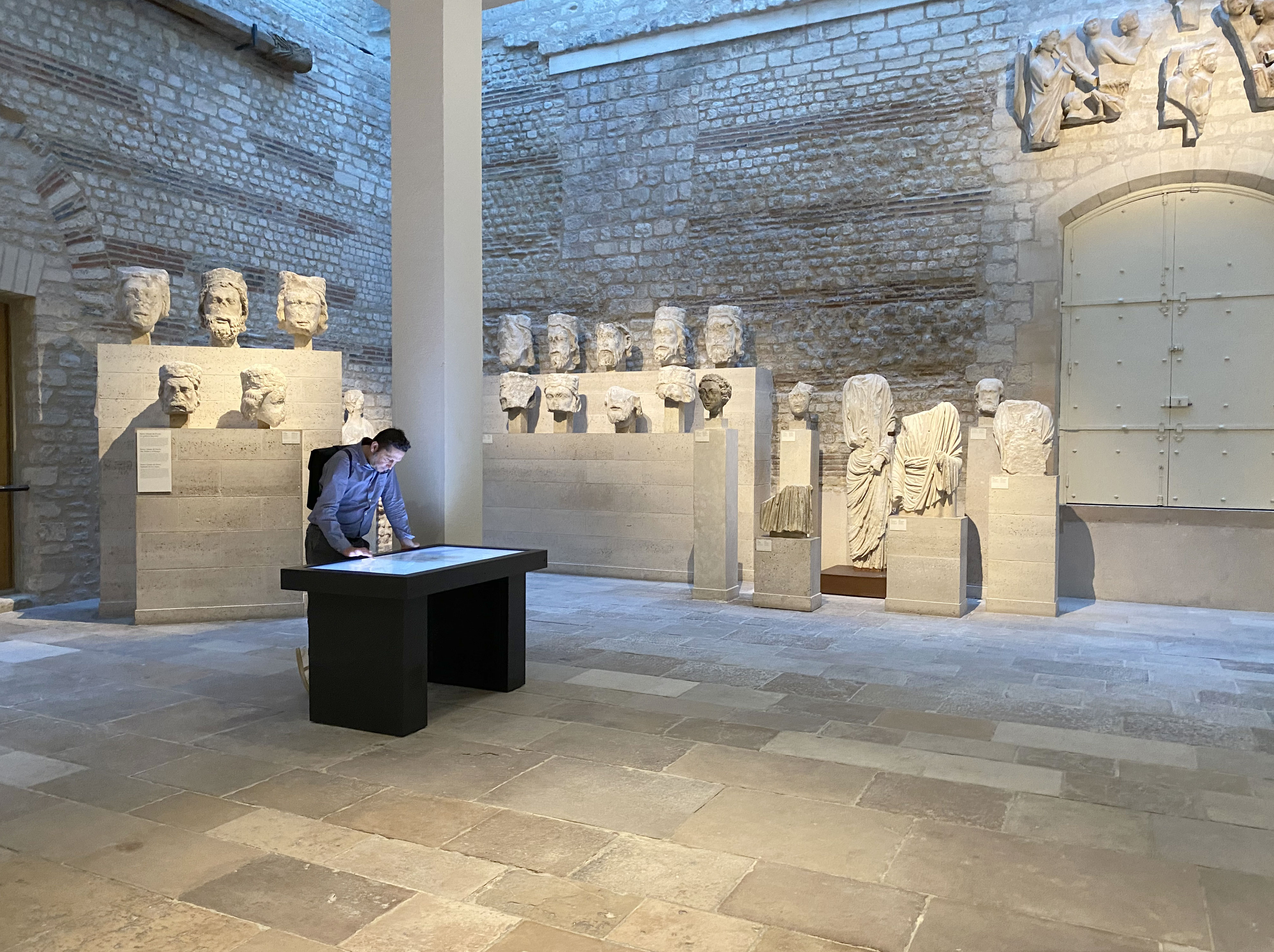 L'image montre un espace intérieur, probablement un musée ou une galerie. On peut y voir plusieurs sculptures en marbre, dont des bustes figurants des visages humains, alignés sur des socles le long des murs. Au centre, une personne se tient à un bureau noir, regardant attentivement quelque chose sur la surface. La lumière naturelle éclaire la pièce, créant une atmosphère calme et contemplative. Les murs en pierre et le sol en dalles ajoutent une note historique à l'environnement.