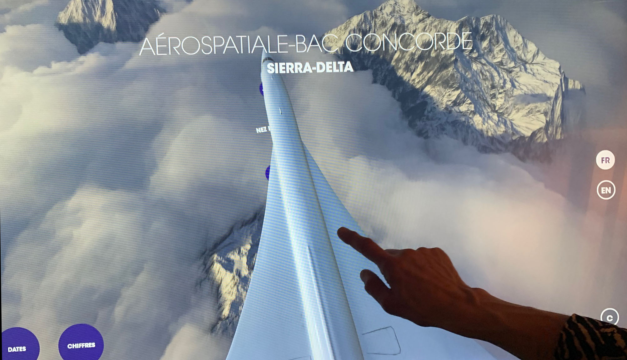 L'image montre une représentation graphique du Concorde, un avion supersonique, vu de l'arrière, s'élevant au dessus des nuages. On aperçoit des montagnes enneigées au loin. Un doigt humain touche l'écran, probablement pour interagir avec une interface qui présente des informations, telles que "Aérospatiale-BAC Concorde" et des éléments en plusieurs langues (FR et EN). Le design semble moderne et technologique.