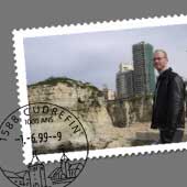 L'image représente un homme se tenant sur un bord, probablement près d'une falaise ou d'une plage. En arrière-plan, on peut voir des bâtiments modernes d'une ville, avec un ciel nuageux. L'image a l'apparence d'une carte postale, avec une bordure blanche et un cachet postal en bas à gauche. L'homme porte une veste en cuir et semble profiter du paysage.