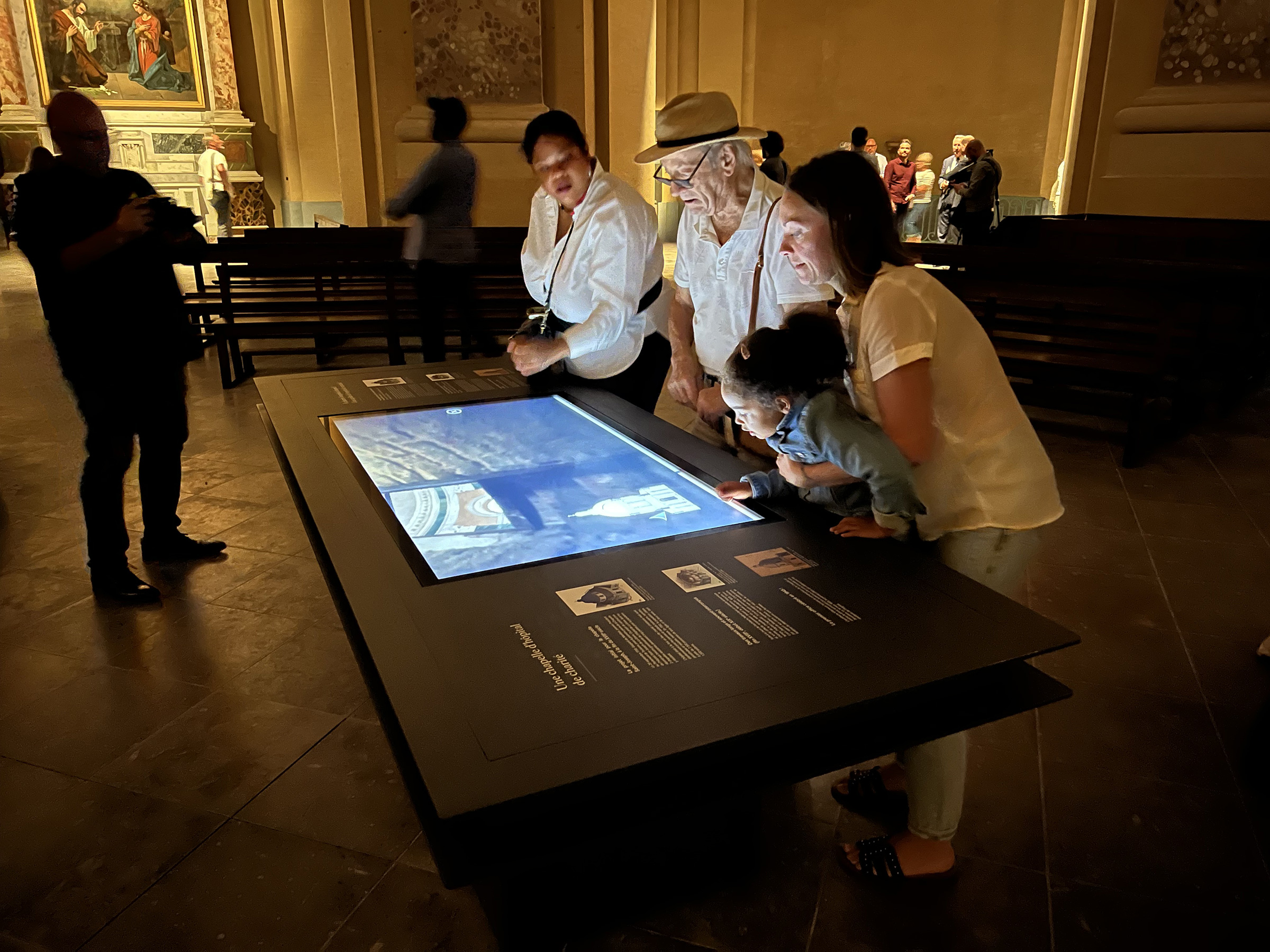 L'image montre un groupe de personnes rassemblées autour d'un grand écran tactile plat, installé dans un espace muséal. On peut voir deux adultes et un enfant regardant attentivement l'écran, qui semble afficher des informations ou des images, probablement liées à une exposition. Un autre adulte, en arrière-plan, observe également la scène. L'environnement est sombre, avec des éclairages chaleureux qui mettent en valeur les détails architecturaux de la salle. L'ambiance semble conviviale et engageante, parfaite pour la découverte culturelle.