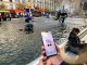 The image depicts a lively urban scene featuring a fountain in a public square. In the background, colorful sculptures are visible, creating a vibrant atmosphere. There are people around, with one person sitting by the fountain, seemingly engaged with their phone. In the foreground, a hand holds a smartphone displaying the app "Ircam Circus." The setting appears to be bright and inviting, with hints of artistic flair in the sculptures and overall environment.