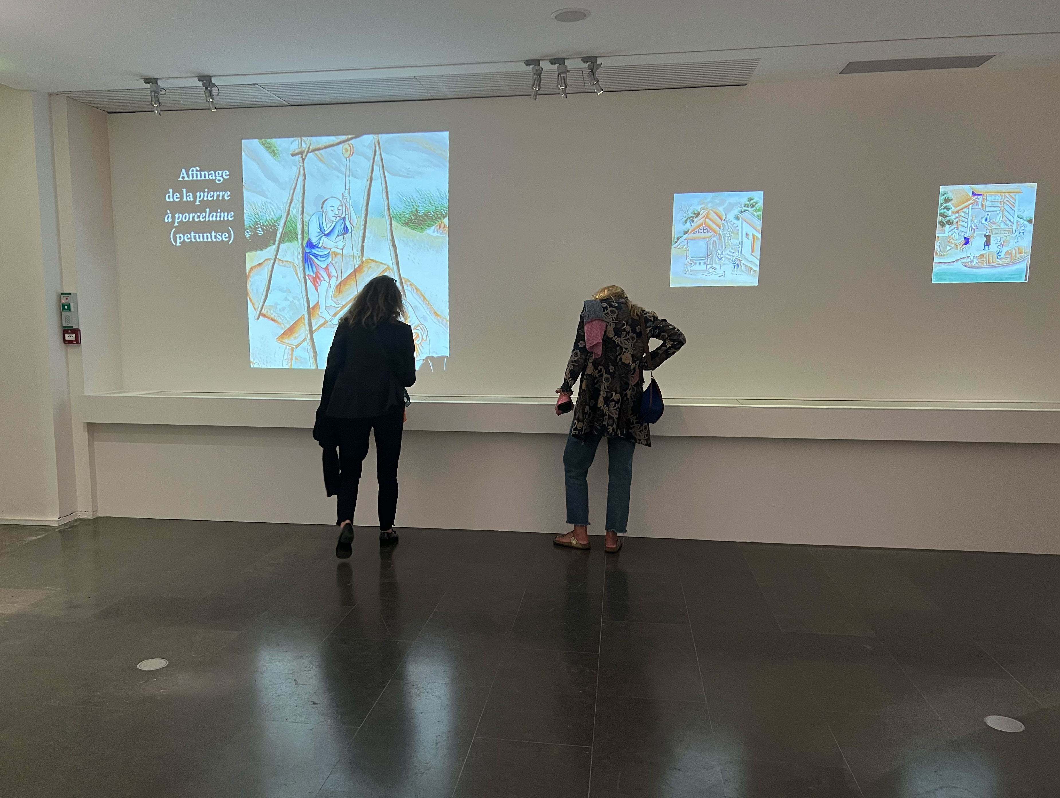 L'image montre un espace d'exposition où deux personnes se tiennent debout devant un mur blanc. Sur ce mur, il y a des projections de plusieurs œuvres, dont l'une semble parler de l'“Afilage de la pierre à procelaine (petuntse)”. Les personnes regardent les projections avec intérêt. L'éclairage est doux et l'environnement semble calme, favorisant une expérience contemplative.