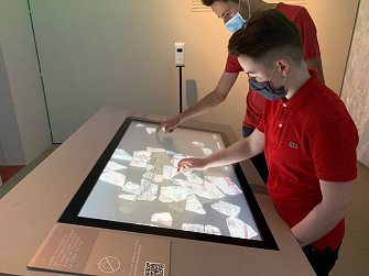 L'image montre deux jeunes garçons interactant avec un écran tactile en exposant des éléments. Ils portent des masques en raison de mesures sanitaires. L'écran affiche de nombreux morceaux colorés, peut-être des artefacts ou des éléments informatiques, et les garçons semblent manipuler ces objets virtuels. L'environnement est moderne et éducatif, suggérant qu'ils se trouvent dans un musée ou une exposition.