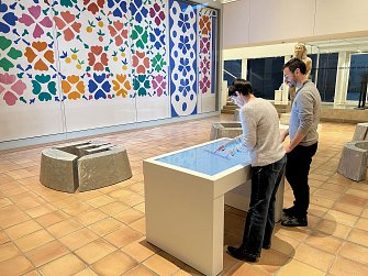L'image montre deux personnes, un homme et une femme, interagissant avec un écran tactile placé sur une table moderne dans un espace muséal. En arrière-plan, une grande œuvre colorée est accrochée au mur, composée de motifs floraux et géométriques. Le sol est carrelé et des statues ou éléments sculpturaux sont visibles autour. L'atmosphère semble contemporaine et artistique.