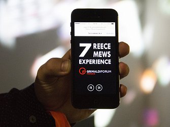 L'image montre un téléphone portable tenu en main, affichant un écran avec des informations sur un événement. Le texte en gros caractères mentionne "7 REECE MEWS EXPERIENCE", et un logo avec le nom "GRIMALDI FORUM MONACO" est visible. Il y a également des options pour se connecter, ainsi que des indications pour choisir la langue (FR pour français et EN pour anglais). L'arrière-plan est flou, avec des formes colorées.