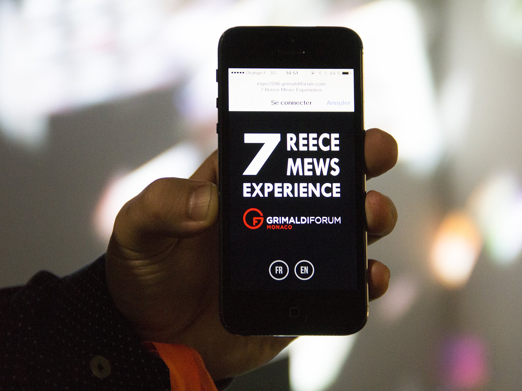 L'image montre un téléphone portable tenu en main, affichant un écran avec des informations sur un événement. Le texte en gros caractères mentionne "7 REECE MEWS EXPERIENCE", et un logo avec le nom "GRIMALDI FORUM MONACO" est visible. Il y a également des options pour se connecter, ainsi que des indications pour choisir la langue (FR pour français et EN pour anglais). L'arrière-plan est flou, avec des formes colorées.