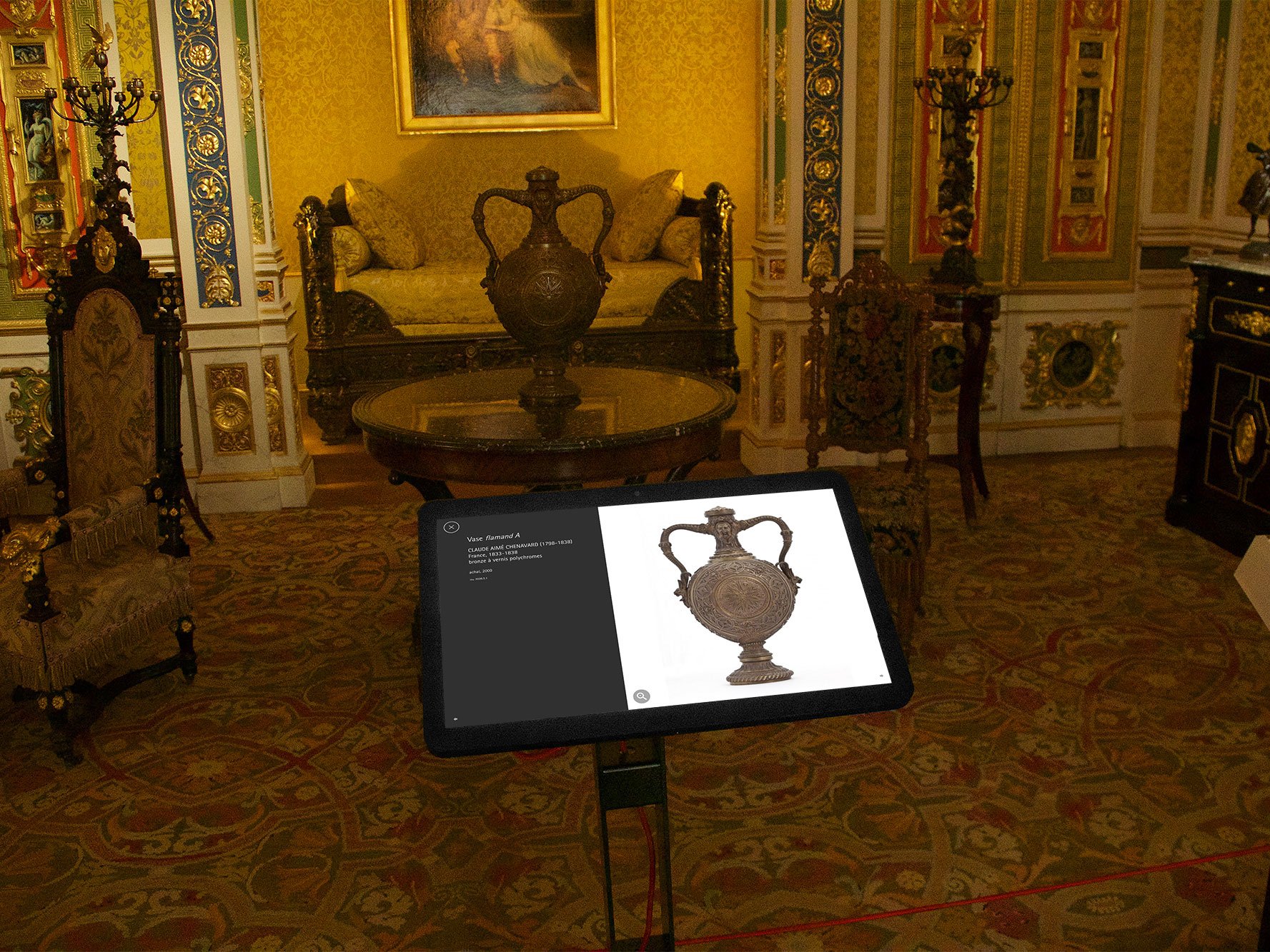 L'image montre une pièce richement décorée, probablement dans un musée ou un palais, avec des détails architecturaux élaborés. Au centre, il y a une table sur laquelle repose une tablette affichant une représentation d'une belle urne ou vase ornée. En arrière-plan, on aperçoit un canapé et des chaises élégantes, toutes avec des motifs raffinés. Le sol est recouvert d'un tapis orné aux couleurs vives. Les murs sont décorés avec des motifs complexes et des œuvres d'art. L'ensemble dégage une atmosphère historique et artistique.